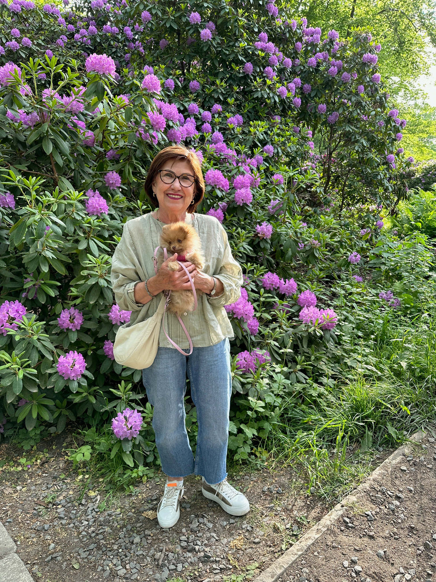 Weibliches Model hält ein Zwergspitzwelpe auf dem Arm, im Hintergrund lilafarbene Rhododendron im Berliner Tiergarten. Der Hund trägt eine vegane schmale Leine in rosa von SUNNY BERLIN